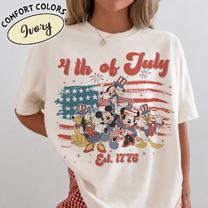 May include: Ivory-colored t-shirt with a patriotic design featuring cartoon characters in red, white, and blue attire. The shirt displays the text "4th of July" and "Est. 1776" with a stylized American flag.