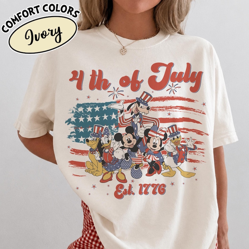 May include: Ivory-colored t-shirt with a patriotic design featuring cartoon characters in red, white, and blue attire. The shirt displays the text "4th of July" and "Est. 1776" with a stylized American flag.