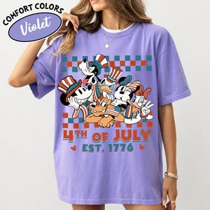May include: A violet t-shirt featuring a graphic of cartoon characters wearing patriotic hats and holding American flags. The design includes a red and teal checkered border, the text 4TH OF JULY, and EST. 1776. A Comfort Colors Violet label is visible.
