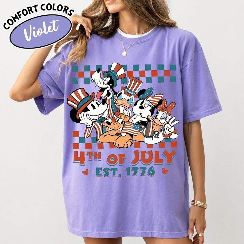 May include: A violet t-shirt featuring a graphic of cartoon characters wearing patriotic hats and holding American flags. The design includes a red and teal checkered border, the text 4TH OF JULY, and EST. 1776. A Comfort Colors Violet label is visible.