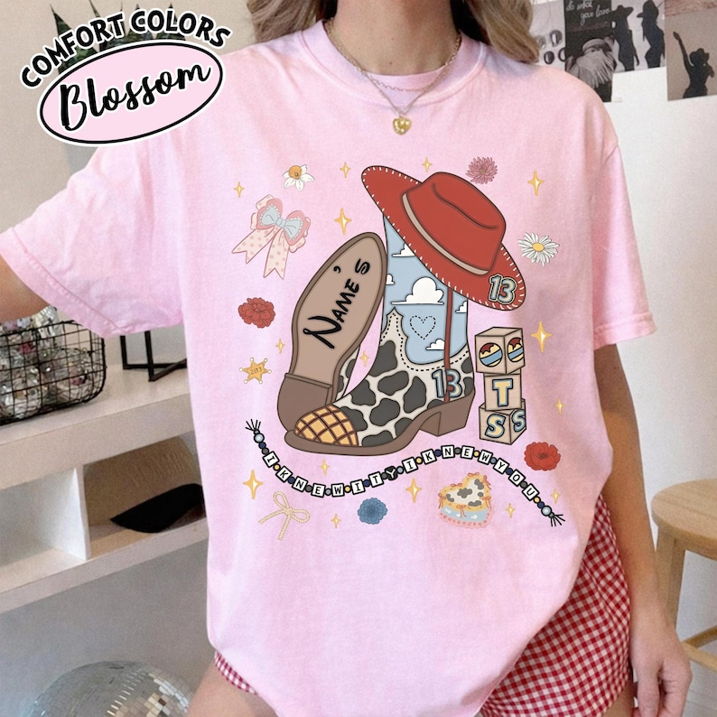 Puede incluir: Una camiseta rosa claro con un estampado gr&aacute;fico de una bota vaquera con manchas de vaca, un sombrero vaquero rojo, bloques con el n&uacute;mero 13 y un collar de cuentas con el texto I KNEW IT I KNEW YOU. La bota incluye el texto NAME'S.
