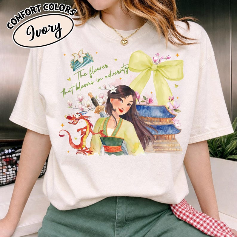 Puede incluir: Camiseta de color marfil con una ilustraci&oacute;n en acuarela de una mujer con un atuendo amarillo y verde, un drag&oacute;n rojo y una pagoda. El texto "The flower that blooms in adversity" est&aacute; impreso en la camiseta.
