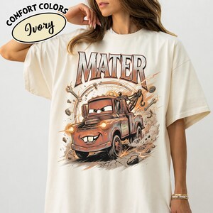Puede incluir: Camiseta de color marfil con una imagen de dibujos animados de Mater de la pel&iacute;cula Cars. El gr&aacute;fico incluye el nombre "MATER" encima del personaje. La camiseta tambi&eacute;n tiene las palabras "COMFORT COLORS" e "Ivory" en un c&iacute;rculo.