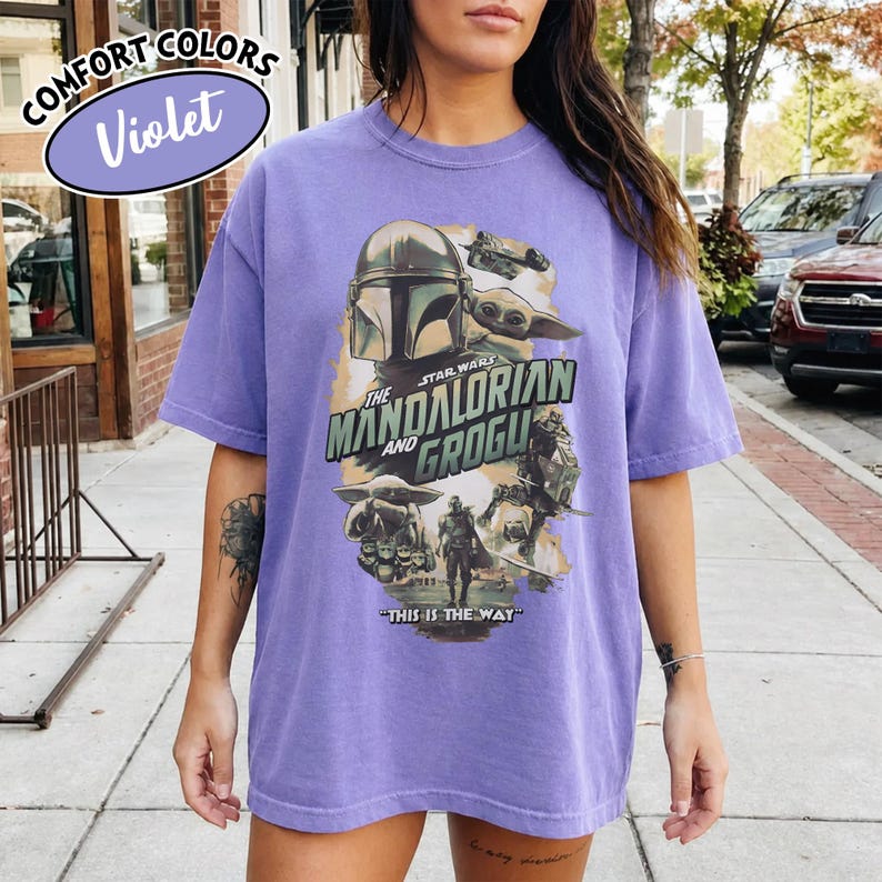 May include: A violet-colored t-shirt featuring a graphic from "Star Wars: The Mandalorian" with the text "The Mandalorian and Grogu" and "This is the Way." The shirt has short sleeves and a relaxed fit.