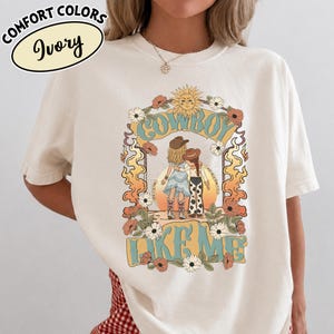 May include: An ivory t-shirt featuring a graphic print of two figures in cowboy hats and boots standing together. The design includes a sun, flowers, and flames with the text "COWBOY LIKE ME" in blue and yellow. A label reads "COMFORT COLORS Ivory".