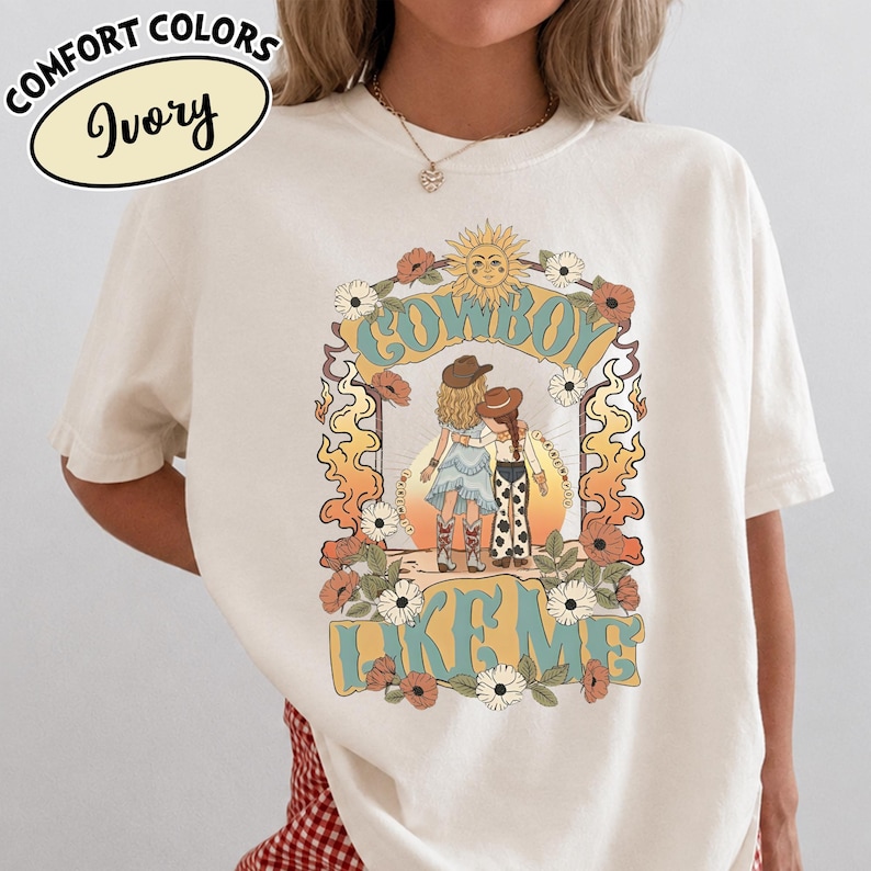 May include: An ivory t-shirt featuring a graphic print of two figures in cowboy hats and boots standing together. The design includes a sun, flowers, and flames with the text "COWBOY LIKE ME" in blue and yellow. A label reads "COMFORT COLORS Ivory".