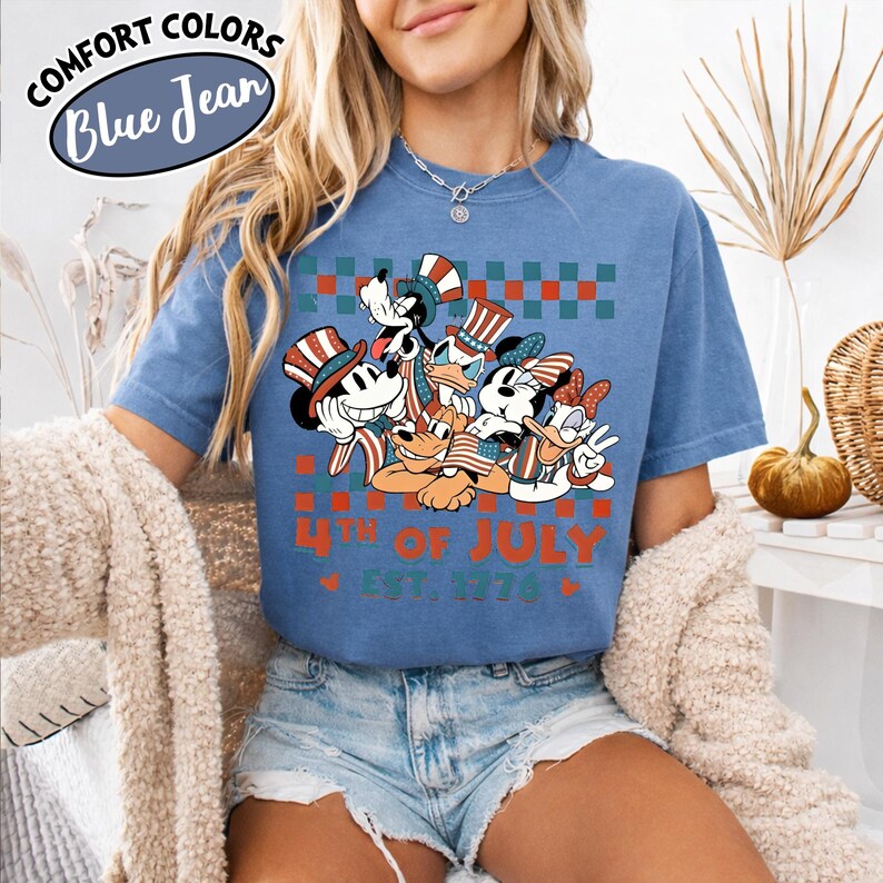 May include: A blue short-sleeve t-shirt featuring a graphic of cartoon characters wearing patriotic hats and holding American flags. The design includes a red and green checkered pattern, the text 4TH OF JULY, and EST. 1776 in orange.