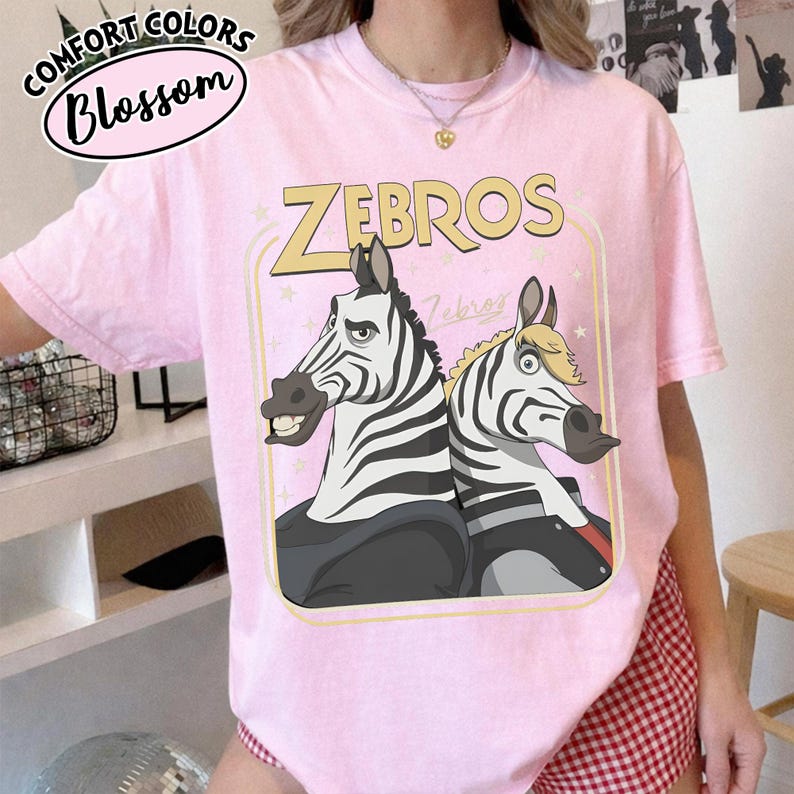 Puede incluir: Camiseta rosa claro con la palabra "ZEBROS" en amarillo, con una ilustraci&oacute;n de dibujos animados de dos cebras. La camiseta tambi&eacute;n tiene el texto "Comfort Colors Blossom" en un dise&ntilde;o circular.