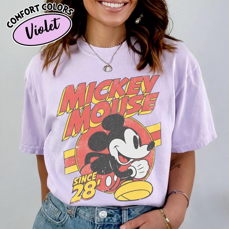 以下が含まれることがあります： ミッキーマウスのヴィンテージグラフィックが特徴のバイオレットカラーのTシャツ。デザインには、赤と黄色で「ミッキーマウス」の文字が、中央にミッキーの画像が描かれています。「Since 28」の文字も表示されています。シャツには「Comfort Colors」と「Violet」の文字が円で囲まれています。