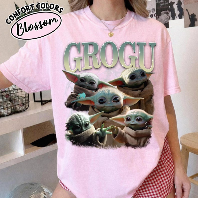May include: A light pink t-shirt featuring the word "GROGU" in a green and silver font, with a graphic of five Grogu characters. The shirt also has the text "COMFORT COLORS" and "Blossom" in a circular design.