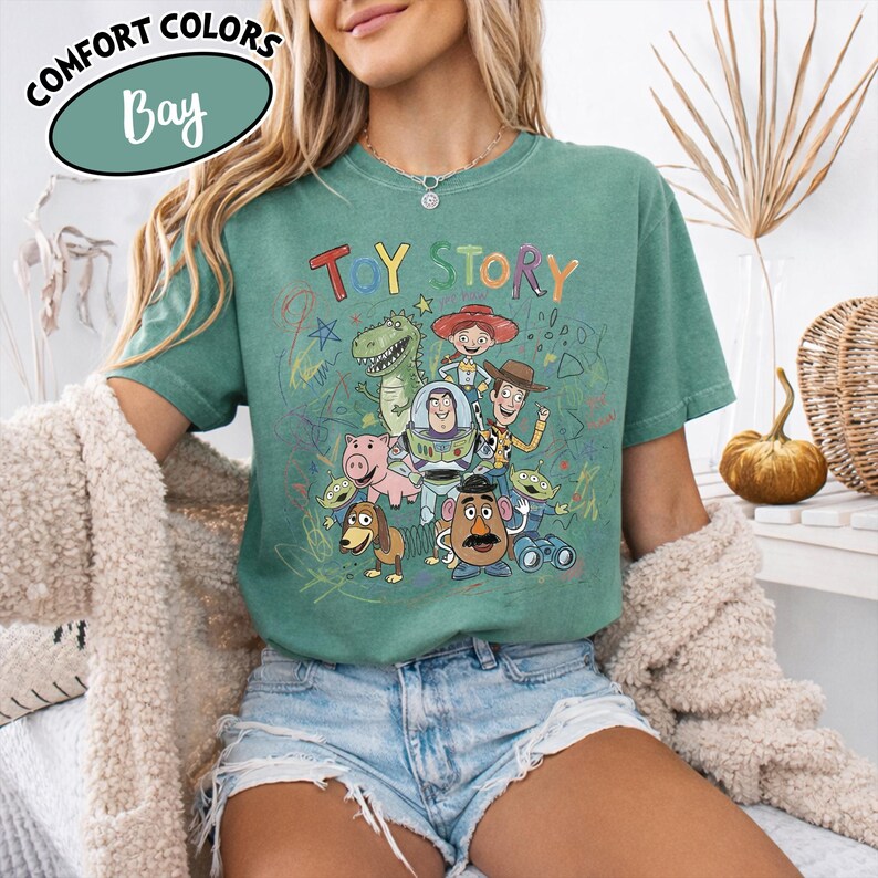 May include: A green t-shirt featuring the words "TOY STORY" in colorful letters, with a graphic of characters from the movie. The shirt has a crew neck and short sleeves. The words "Comfort Colors" and "Bay" are in a circle in the upper left corner.