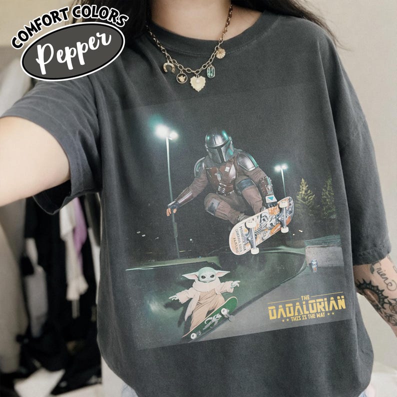 May include: Dark grey Comfort Colors t-shirt featuring a graphic of The Mandalorian and Grogu skateboarding in a skate park. The text "THE DADALORIAN...THIS IS THE WAY" is printed below the image. The shirt has the text "COMFORT COLORS Pepper" in a white oval.