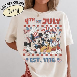 May include: An ivory t-shirt featuring a graphic print with the text 4TH OF JULY and EST. 1776. The design includes cartoon characters wearing patriotic hats, a small American flag, and fireworks against a red and blue checkered background.