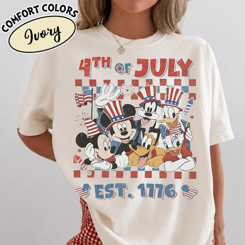 May include: An ivory t-shirt featuring a graphic print with the text 4TH OF JULY and EST. 1776. The design includes cartoon characters wearing patriotic hats, a small American flag, and fireworks against a red and blue checkered background.