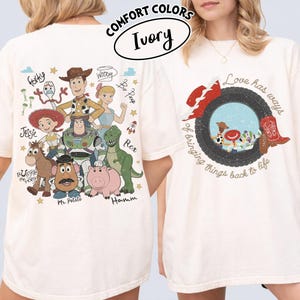 May include: An ivory t-shirt featuring a graphic print of Toy Story characters including Woody, Buzz Lightyear, Jessie, Rex, Hamm, Bullseye, Forky, and Bo Peep on the back, with a circular graphic and the text Love has ways of bringing things back to life on the front.