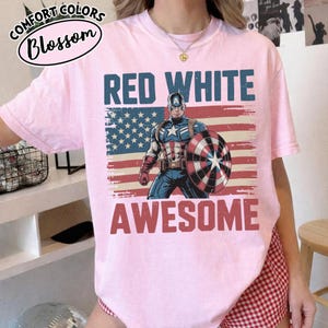 K&ouml;nnte beinhalten: Hellrosa T-Shirt mit den Worten "RED WHITE AWESOME" in Rot und Blau, mit einer Illustration von Captain America vor einer amerikanischen Flagge. Das Shirt hat das Comfort Colors Blossom Logo.