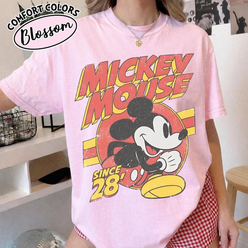 以下が含まれることがあります： ミッキーマウスのヴィンテージグラフィックが特徴のライトピンクのTシャツ。「ミッキーマウス」の文字は黄色と赤で、「Since 28」の文字も含まれています。丸首で半袖のTシャツです。