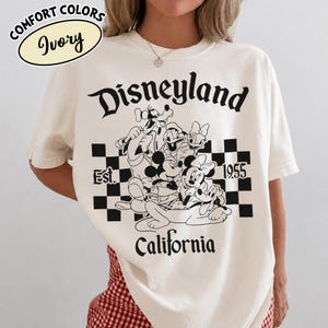 May include: Ivory-colored t-shirt with a Disneyland graphic. The design features Mickey Mouse, Minnie Mouse, Goofy, Donald Duck, and Pluto in a black and white checkered box. The text "Disneyland California Est. 1955" is also printed on the shirt.