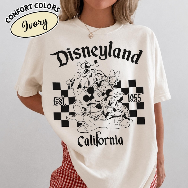 May include: Ivory-colored t-shirt with a Disneyland graphic. The design features Mickey Mouse, Minnie Mouse, Goofy, Donald Duck, and Pluto in a black and white checkered box. The text "Disneyland California Est. 1955" is also printed on the shirt.