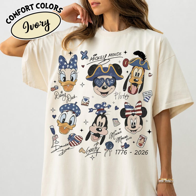 May include: An ivory t-shirt featuring a graphic print of Disney characters including Mickey Mouse, Minnie Mouse, Donald Duck, Daisy Duck, Goofy, and Pluto. The design includes patriotic stars and stripes motifs, the text 1776 - 2026, and character names written in script. The shirt is labelled Comfort Colors Ivory.