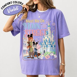May include: Violet Comfort Colors t-shirt featuring a graphic of cartoon characters and a castle with the text "Meet Me On Main Street". The design includes a fox and a rabbit, with fireworks and other details.