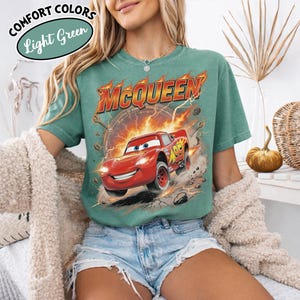 May include: Light green t-shirt featuring a graphic of Lightning McQueen from the movie Cars. The design includes the name "McQueen" in orange and red, with flames and a racing number. The shirt also has the text "Comfort Colors" and "Light Green".