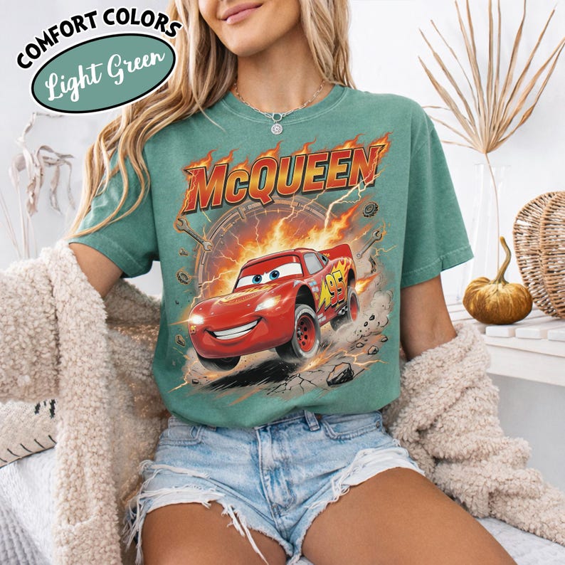 May include: Light green t-shirt featuring a graphic of Lightning McQueen from the movie Cars. The design includes the name "McQueen" in orange and red, with flames and a racing number. The shirt also has the text "Comfort Colors" and "Light Green".