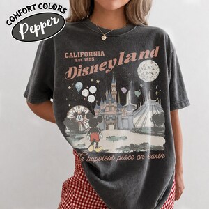 May include: Dark gray t-shirt with a Disneyland graphic. The design features a castle, Mickey Mouse, a Ferris wheel, and the text "California Disneyland Est. 1955" and "the happiest place on earth". The shirt has a vintage, distressed look.