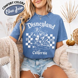 May include: Blue jean colored t-shirt with a white graphic of Disneyland characters and text. The graphic includes the words "Disneyland", "Est. 1955", and "California". The shirt has short sleeves and a crew neck.