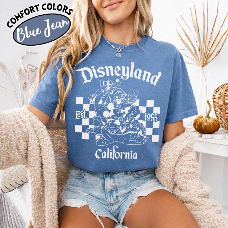 May include: Blue jean colored t-shirt with a white graphic of Disneyland characters and text. The graphic includes the words "Disneyland", "Est. 1955", and "California". The shirt has short sleeves and a crew neck.
