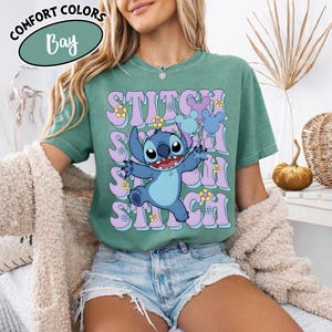Puede incluir: Una camiseta verde con el personaje Stitch de Lilo & Stitch. El dise&ntilde;o incluye la palabra "Stitch" repetida en una fuente retro con flores y globos de Mickey Mouse. La camiseta tambi&eacute;n tiene las palabras "Comfort Colors" y "Bay" en un c&iacute;rculo.