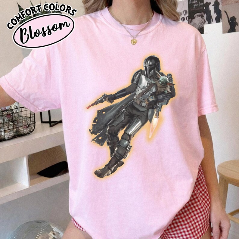 Puede incluir: Una camiseta de manga corta de color rosa claro con un estampado gr&aacute;fico de un personaje con armadura plateada que sostiene a una peque&ntilde;a criatura verde. El personaje lleva casco y capa, y sostiene un bl&aacute;ster. El texto Comfort Colors Blossom es visible.