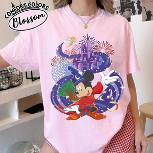 Może przedstawiać: Jasnor&oacute;żowa koszulka z grafiką Myszki Miki w stroju czarodzieja, z zamkiem, fajerwerkami i wr&oacute;żką. Koszulka ma napis "Comfort Colors Blossom" w okrągłym wzorze.