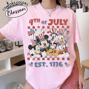 May include: A light pink short-sleeve t-shirt featuring a graphic print of cartoon characters wearing patriotic hats. The text reads 4TH OF JULY and EST. 1776. The design includes a red and blue checkered background with stars and stripes motifs.