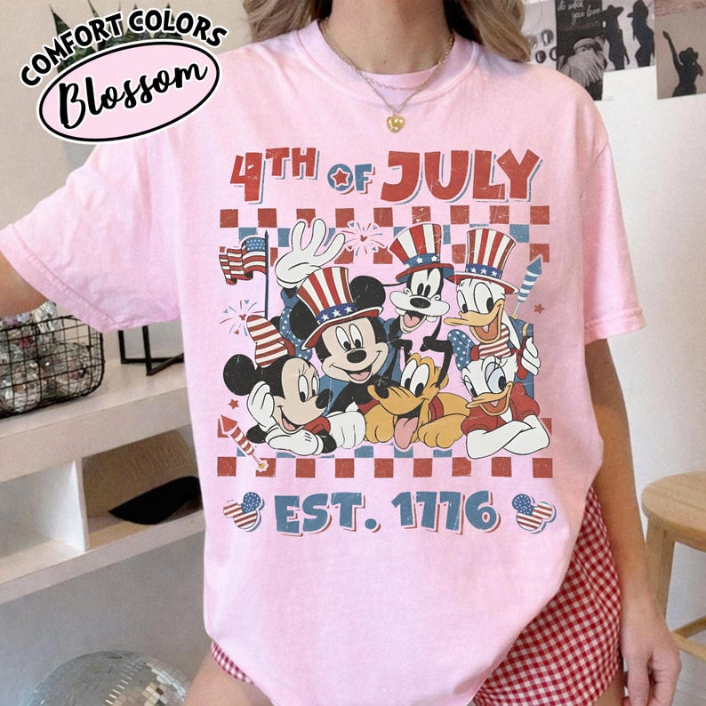 May include: A light pink short-sleeve t-shirt featuring a graphic print of cartoon characters wearing patriotic hats. The text reads 4TH OF JULY and EST. 1776. The design includes a red and blue checkered background with stars and stripes motifs.