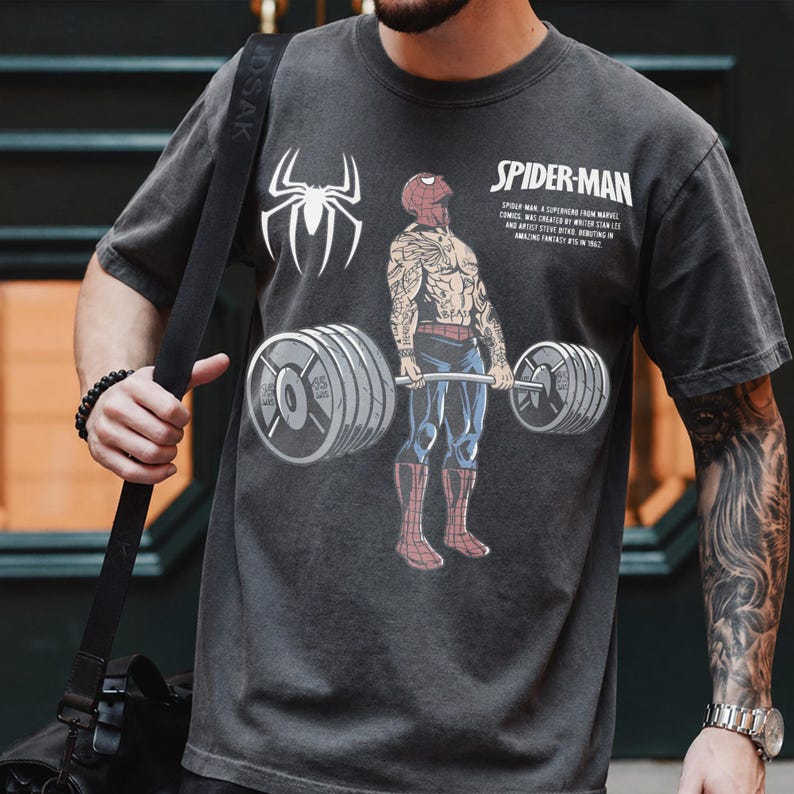 May include: Dark gray t-shirt featuring a Spider-Man graphic lifting weights. The design includes a white spider symbol and the text "SPIDER-MAN". The shirt has a vintage, worn look.