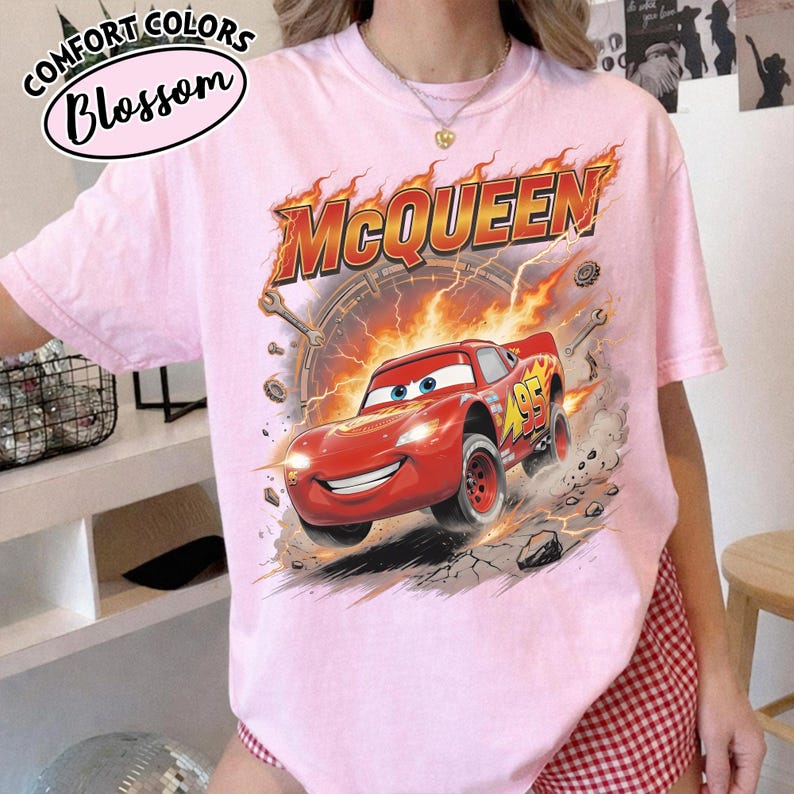 May include: Pink t-shirt with a graphic of Lightning McQueen from the movie Cars. The graphic features the car with flames and the word "McQueen". The shirt also has the words "Comfort Colors Blossom" in a circle.