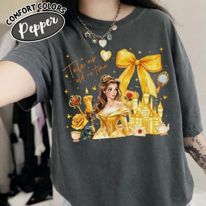 Puede incluir: Camiseta Comfort Colors gris oscuro con una ilustraci&oacute;n colorida de una mujer con un vestido amarillo, un castillo y el texto "Tale as old as time". Un gran lazo amarillo en la parte superior.
