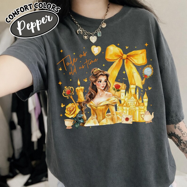 Puede incluir: Camiseta Comfort Colors gris oscuro con una ilustraci&oacute;n colorida de una mujer con un vestido amarillo, un castillo y el texto "Tale as old as time". Un gran lazo amarillo en la parte superior.
