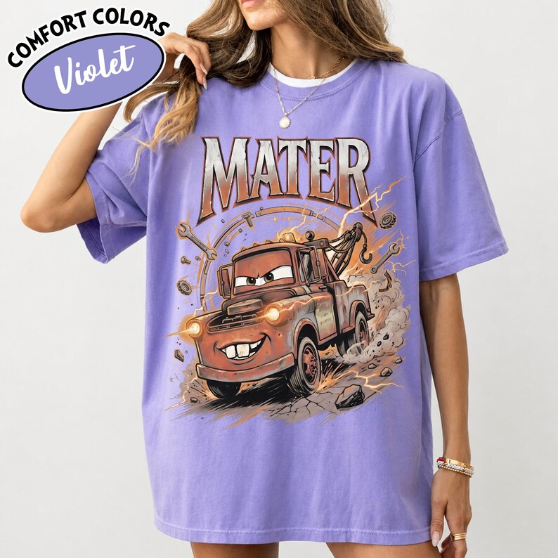Puede incluir: Camiseta violeta Comfort Colors con una imagen de dibujos animados de Mate de la pel&iacute;cula Cars. El gr&aacute;fico incluye el nombre "MATER" en naranja y una gr&uacute;a Mate sonriente con herramientas y rayos. La camiseta tiene un corte holgado.
