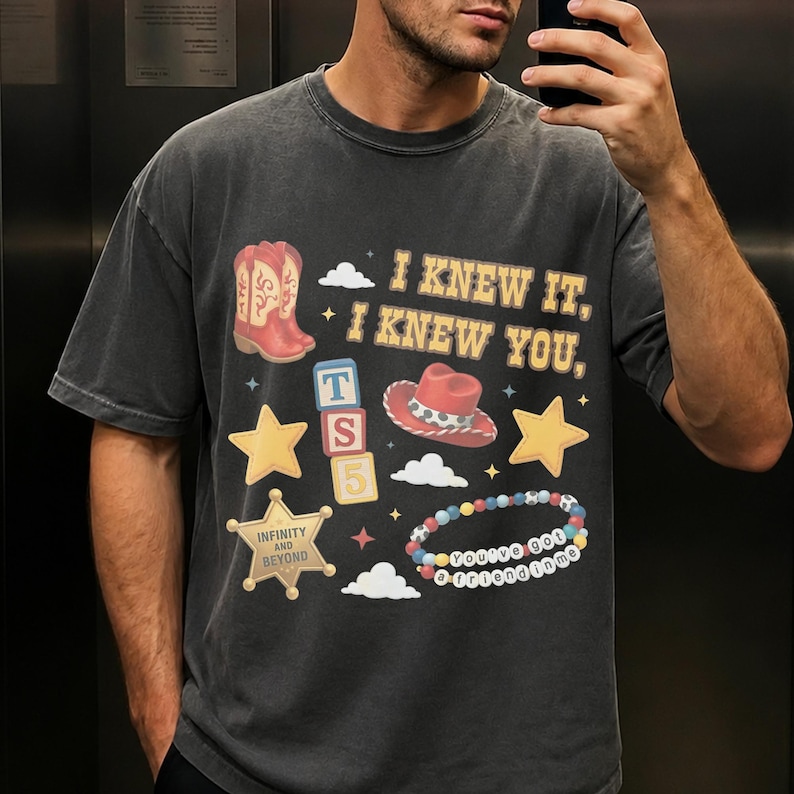 May include: A charcoal gray short-sleeve t-shirt featuring a graphic print with the text I KNEW IT, I KNEW YOU, a red cowboy boot, a red cowboy hat, yellow stars, a sheriff badge with INFINITY AND BEYOND, alphabet blocks, and a beaded bracelet.