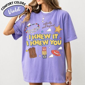 May include: A violet short-sleeved t-shirt featuring a graphic print with the text "I KNEW IT I KNEW YOU", "infinity and beyond", and "YOU'VE GOT A FRIEND IN ME". The design includes illustrations of cowboy boots, a red hat, and alphabet blocks.