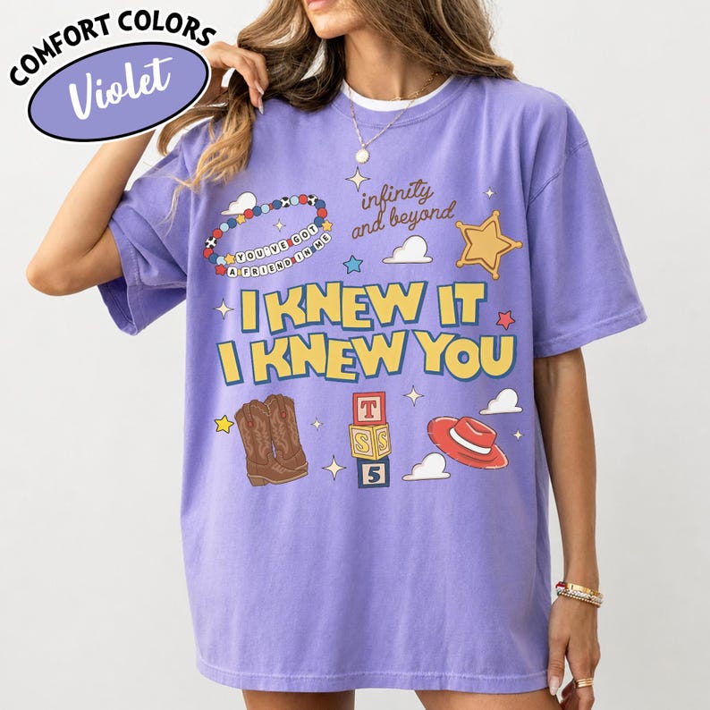 May include: A violet short-sleeved t-shirt featuring a graphic print with the text "I KNEW IT I KNEW YOU", "infinity and beyond", and "YOU'VE GOT A FRIEND IN ME". The design includes illustrations of cowboy boots, a red hat, and alphabet blocks.