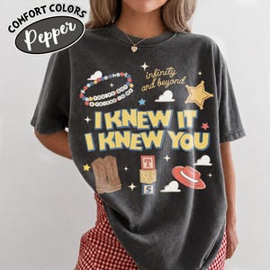 May include: A charcoal gray short-sleeve t-shirt featuring a graphic print with the text "I KNEW IT I KNEW YOU," "infinity and beyond," and "YOU'VE GOT A FRIEND IN ME." The design includes illustrations of a beaded bracelet, a sheriff star, cowboy boots, alphabet blocks, and a red hat.