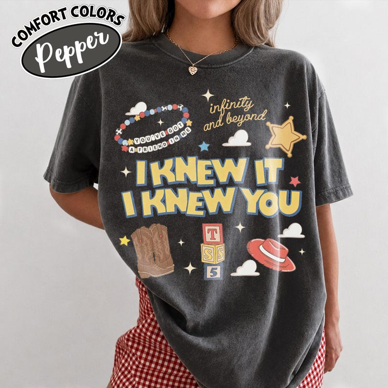May include: A charcoal gray short-sleeve t-shirt featuring a graphic print with the text "I KNEW IT I KNEW YOU," "infinity and beyond," and "YOU'VE GOT A FRIEND IN ME." The design includes illustrations of a beaded bracelet, a sheriff star, cowboy boots, alphabet blocks, and a red hat.