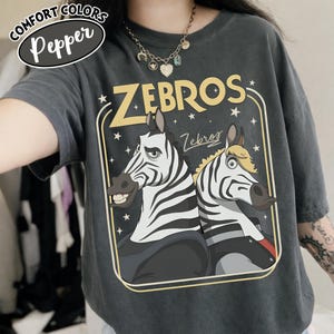Puede incluir: Camiseta gris oscuro Comfort Colors con un gr&aacute;fico de dos cebras y la palabra "ZEBROS" en amarillo. Las cebras tienen rayas blancas y negras y est&aacute;n sobre un fondo oscuro con estrellas.