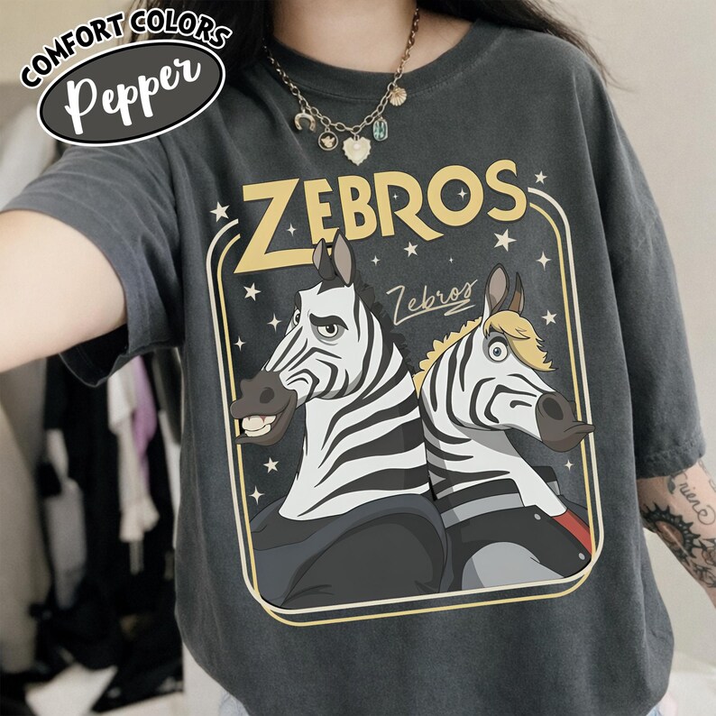 Puede incluir: Camiseta gris oscuro Comfort Colors con un gr&aacute;fico de dos cebras y la palabra "ZEBROS" en amarillo. Las cebras tienen rayas blancas y negras y est&aacute;n sobre un fondo oscuro con estrellas.