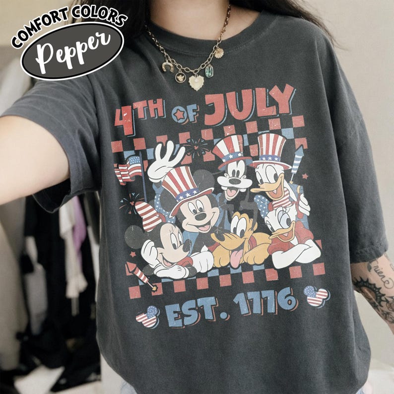 May include: A charcoal gray t-shirt featuring a graphic print of cartoon characters wearing patriotic hats. The text reads 4TH OF JULY and EST. 1776. The design includes a checkered background, fireworks, and small American flag motifs in red, white, and blue.