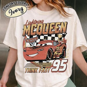 May include: Ivory t-shirt with a distressed graphic of Lightning McQueen from the Cars film. The design includes the text "Lightning McQueen", "95", and "Think Fast". The shirt has a classic crew neck and short sleeves.