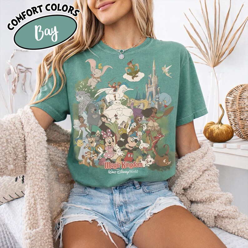 Op de afbeelding: Groen t-shirt met een kleurrijke afbeelding van Disney-personages, waaronder Mickey Mouse en Peter Pan. Het shirt heeft de tekst "Magic Kingdom" en "Walt Disney World". Het shirt heeft ook de tekst "Comfort Colors Bay".